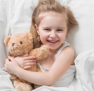 Lovely little blonde girl holding her favorite brown teddy, lying in bed, smiling widely