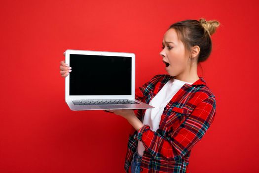 photo of Beautiful shocked amazed young woman holding laptop wearing red shirt looking at monitor display isolated on red background. Mock up