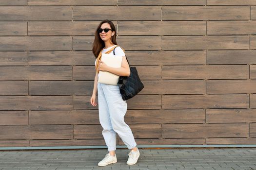 Full length photo shot of young charming smiling happy beautifyl brunet woman going in the street near brown wall in white t-shirt and light blue jeans white sneakers with black female bag holding computer laptop and listening to music with wireless headphones.