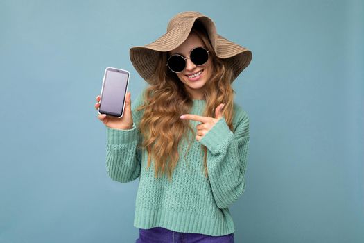 Photo of beautiful smiling young woman good looking wearing casual stylish outfit standing isolated on background with copy space holding smartphone showing phone in hand with empty screen display for mockup pointing at gadjet looking at camera.
