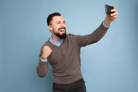 Photo of positive handsome young brunette unshaven man with beard wearing casual grey sweater and blue shirt isolated on pink background wall holding smartphone taking selfie photo looking at mobile phone screen display and showing fist.