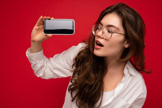 photo of sexy beautiful young brunette woman wearing white shirt and optical glasses isolated over red background holding in hand and showing mobile phone with empty screen for cutout looking at gadjet screen.