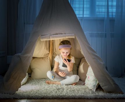 Cheerful little girl in pajamas sitting in cozy teepee tent in her room