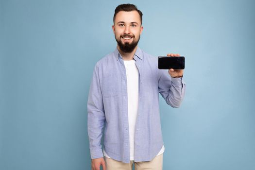 Photo of serious handsome good looking young brunette unshaven man with beard wearing casual white t-shirt and blue shirt poising isolated on blue background with empty space holding in hand mobile with empty screen looking at camera. cutout