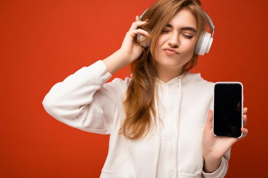 Attractive emotional young blond woman wearing white hoodie isolated over colorful background wall holding and showing mobile phone with empty screen for cutout and wearing bluetooth headphones listening to pop music and enjoying.