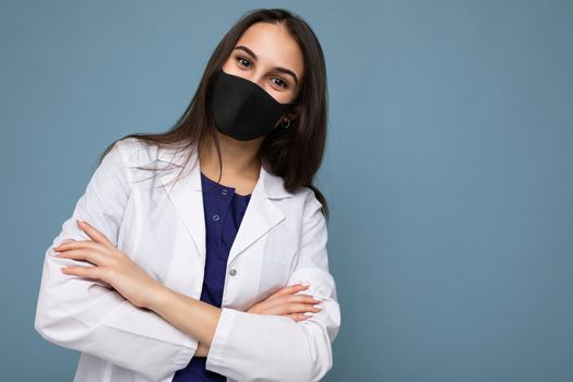 Happy brunette woman in virus protective mask on face against coronavirus and white medical coat isolated on blue background. Empty space