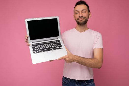 Handsome smiling brunet man holding laptop computer looking at camera in t-shirt on isolated pink background.
