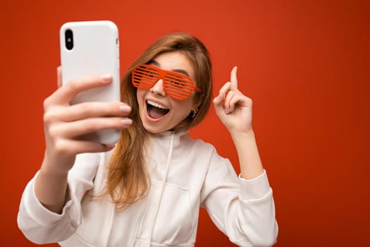 Closeup portrait of attractive positive surprised young blonde woman wearing stylish white hoodie and funny colorful glasses standing isolated over colourful background holding and using mobile phone taking selfie photo looking at gadjet and pointing finger up. copy space