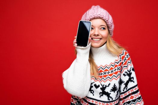 Photo of attractive smiling young blonde woman wearing warm knitted hat and winter warm sweater standing isolated over red background showing smartphone with empty screen for cutout looking to the side.