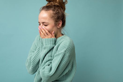 Portrait of young laughing positive nice winsome blond woman with sincere emotions wearing trendy blue sweater isolated over blue background with free space and covering mouth.