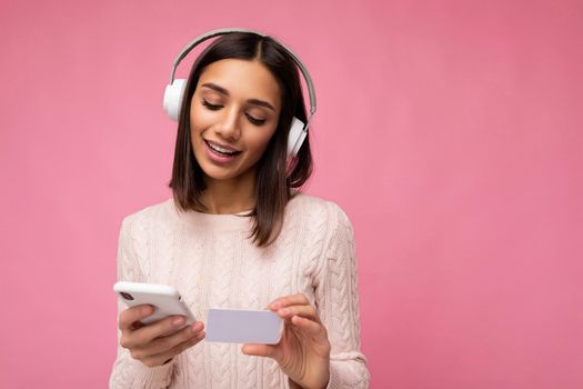 Photo of beautiful positive young brunette woman wearing pink casual sweater isolated over pink background wall wearing white bluetooth wireless headphones and listening to music and using mobile phone making payment online through credit card looking down.