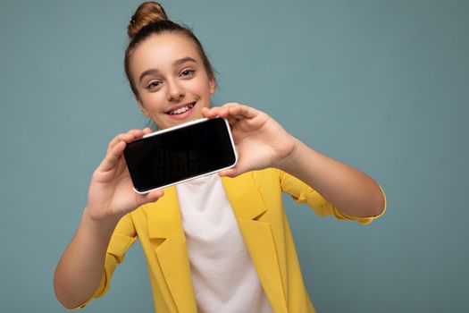 Photo of beautiful smiling teen girl good looking wearing casual stylish outfit standing isolated on background with copy space holding smartphone showing phone in hand with empty screen display for mockup looking at camera.