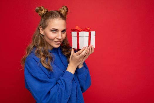 Photo shot of attractive happy sexy adult blond woman isolated over red background wall wearing blue trendy hoodie holding gift box and looking at camera.