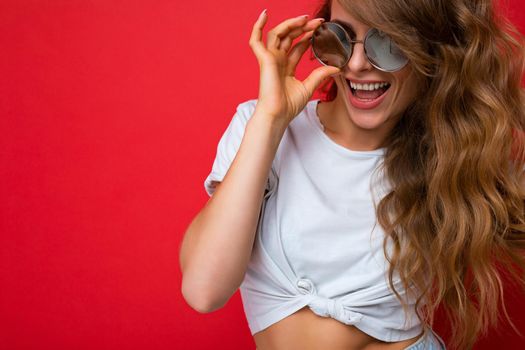 Closeup shot of beautiful smiling young dark blonde curly female person isolated over red background wall wearing casual white t-shirt and stylish sunglasses looking at camera. free space