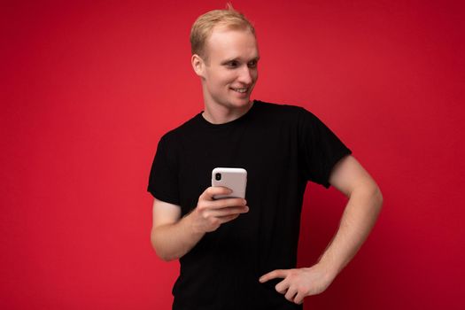 Photo shot of handsome positive good looking young man wearing casual stylish outfit poising isolated on background with empty space holding in hand and using mobile phone messaging sms looking to the side.