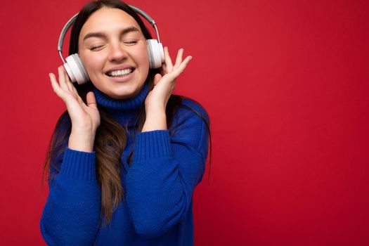 Photo of beautiful happy smiling young brunette woman wearing blue sweater isolated over red background wall wearing white bluetooth headphones listening to cool music enjoying with close eyes. copy space