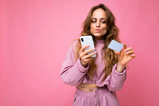 Beautiful sexy positive young blonde curly woman wearing pink clothes isolated over pink background using mobile phone paying online shopping through credit card looking at smartphone display.