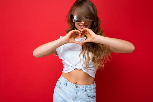 Photo shot of beautiful young dark blonde woman wearing casual clothes and stylish sunglasses isolated over colorful background looking at camera and showing heart gesture.