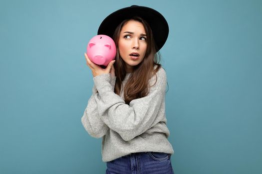 Portrait of positive shocked amazed young beautiful pretty brunette woman wearing gray pullover and black hat isolated over blue background with free space and holding pink piggy box. Money box concept.