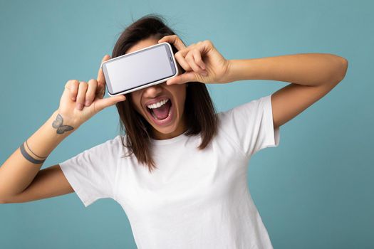 Photo of pretty positive young brunette woman good looking wearing white t-shirt standing isolated on blue background with copy space holding smartphone showing phone in hand with empty screen screen for mockup and having fun.