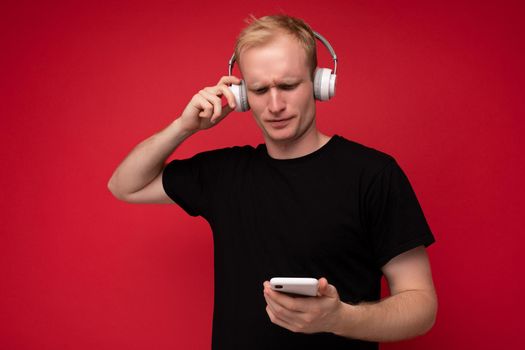 Upset thoughtful handsome blonde young man wearing black t-shirt and white headphones standing isolated over red background with copy space holding smartphone and communicating online listening to music and having fun looking at gadjet screen and don't understanding.