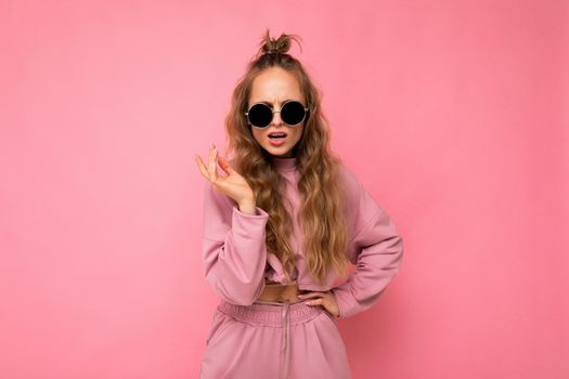 Fascinating dissatisfied adult blonde curly woman isolated over pink background wall wearing casual pink sport clothes and stylish sunglasses looking at camera.