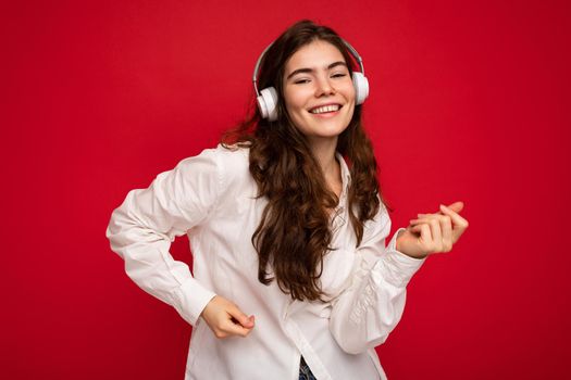 Attractive happy smiling young brunette female person wearing white shirt and optical glasses isolated over red background wearing white wireless bluetooth earphones listening to music and dancing looking at camera.