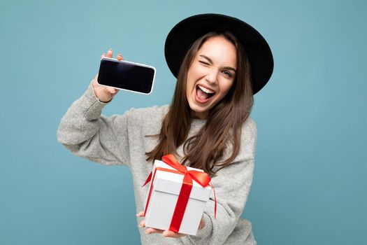 Attractive happy joyful young brunette woman isolated over blue background wall wearing fashionable black hat and casual grey sweater holding gift box showing mobile phone screen display for mockup and looking at camera and winking.