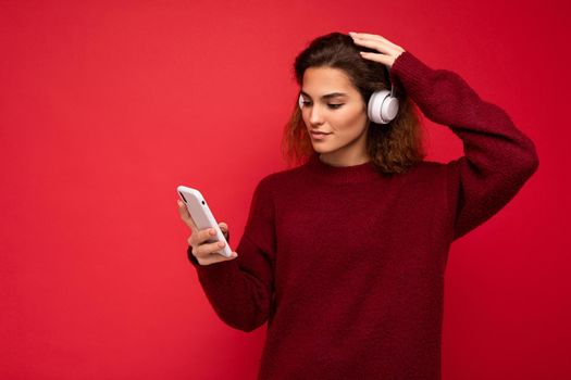 Shot of beautiful young brunette curly woman wearing dark red sweater isolated over red background wall wearing white wireless headphones and listening to music and using mobile phone surfing on the internet looking at smartphone display.