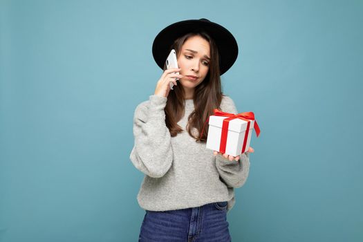 Photo of beautiful unhappy young brunette woman isolated over blue background wall wearing black hat and grey sweater holding gift box talking on mobile phone and looking to the side.