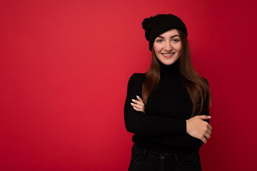 Beautiful positive young brunette woman wearing black sweater and black hat standing isolated over red background looking at camera and smiling keeping hands crossing.