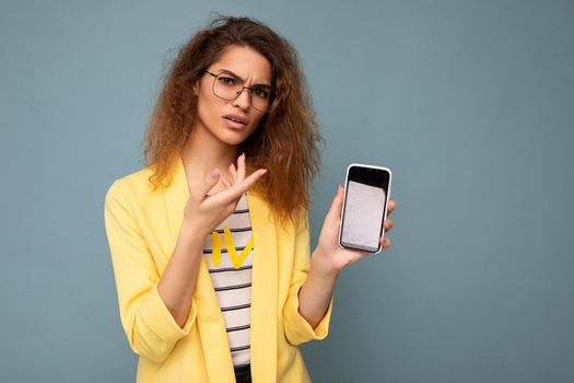 Dissatisfied angry attractive young woman with curly dark blond hair wearing yellow jacket and optical glasses isolated on background holding and showing mobile phone with empty space for cutout looking at camera.