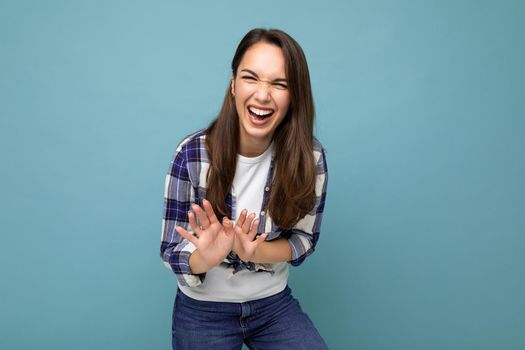 Young positive smiling beautiful brunette woman with sincere emotions wearing trendy check shirt standing isolated on blue background with empty space and showing stop gesture and laughing.