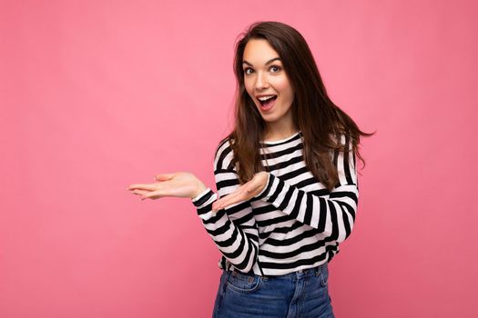 Portrait photo of young happy positive beautiful brunette woman with sincere emotions wearing casual striped pullover isolated on pink background with copy space and pointing hands at free space for text.