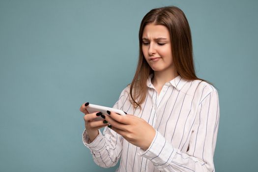 Concentrated attractive young blonde woman wearing casual white shirt isolated over blue background wall holding smartphone and playing online games looking at gadjet screen.