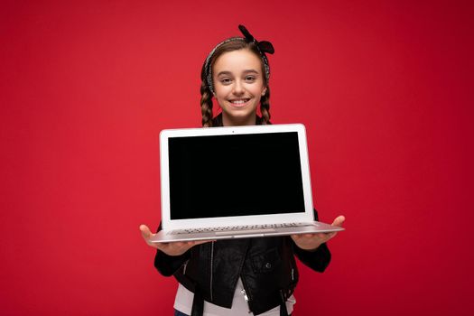 Photo of beautiful happy smiling girl with brunet pigtails holding computer laptop wearing black jacket and bandana isolated over red wall background looking at camera. Mock up, free space for advertising