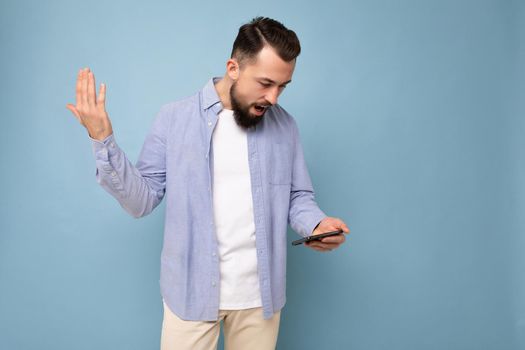 Photo shot of dissatisfied handsome bearded good looking young man wearing casual stylish outfit poising isolated on background with empty space holding in hand and using mobile phone messaging sms looking at smartphone display screen.