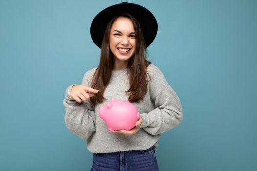 Portrait of happy positive smiling young beautiful brunette woman wearing stylish gray sweater and black hat isolated over blue background with free space and holding pink penny bank. Money box concept.
