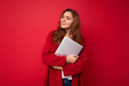 shot of mysterious Beautiful young dark hair curly woman holding close laptop wearing red cardigan and white blouse looking to the side isolated over red background.