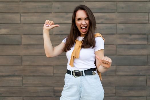 Photo of young happy joyful attractive brunette woman with sincere emotions wearing stylish outfit standing in the street near brown wall and pointing at herself. Lifestyle concept.
