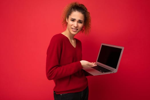 Close-up portrait of beautiful amazed surprised astonished dark blond woman holding laptop computer looking at camera typing on keyboard wearing red sweater isolated over red wall background.