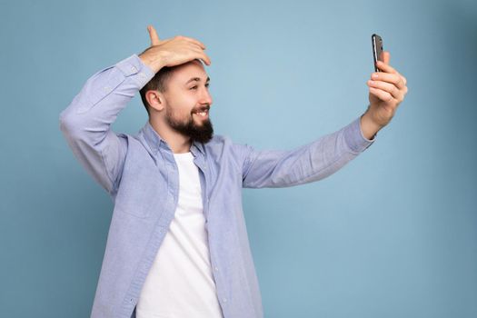 Handsome young brunet bearded man wearing casual stylish clothes standing isolated over blue background wall holding smartphone taking selfie photo looking at mobile phone screen display.