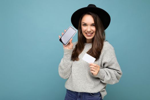 Beautiful smiling young brunette female person wearing black hat and grey sweater isolated over blue background holding credit card and mobile phone with empty display for mockup looking at camera.