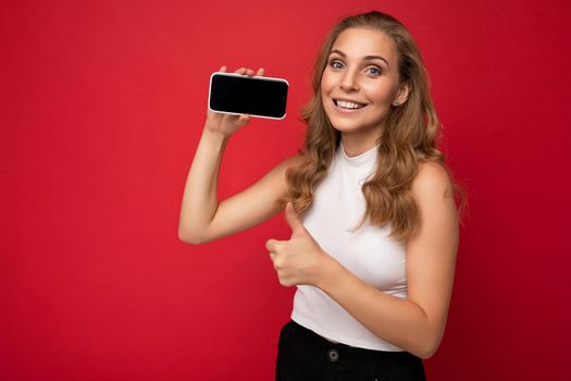 Smiling beautiful young blonde woman wearing white t-shirt isolated on red background with copy space holding smartphone showing phone in hand with empty screen for mockup looking at camera and giving thumbs up gesture.