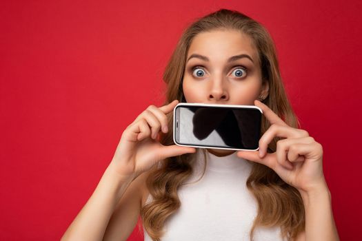 Amazed surprised attractive young blonde female person wearing white t-shirt isolated on red background with copy space holding smartphone showing phone in hand with empty screen for cutout looking at camera.