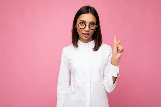 Attractive sexy young brunette woman wearing everyday stylish clothes and modern optical glasses isolated on colorful background wall looking at camera showing one finger.