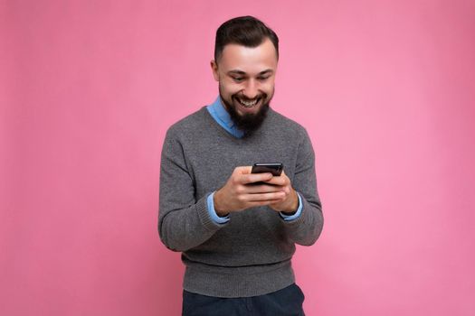 Positive smiling handsome good looking brunet bearded young man wearing grey sweater and blue shirt isolated on pink background with empty space holding in hand and using mobile phone communicating online looking at gadjet screen.