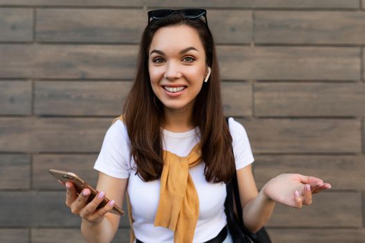 Photo of attractive smiling young woman wearing casual clothes standing in the street holding mobile looking at camera.Copy space