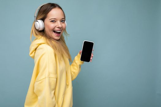 Side-profile photo of beautiful happy smiling young woman wearing stylish casual outfit isolated on background wall holding and showing mobile phone with empty display for mockup wearing white bluetooth headphones listening to music and having fun looking at camera and winking.