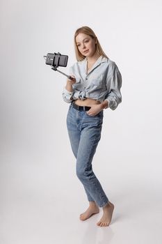 happy female making self portrait with smartphone attached to selfie stick pole. young pretty blond girl with long hair smiling and using cell phone isolated on white background. modern communication